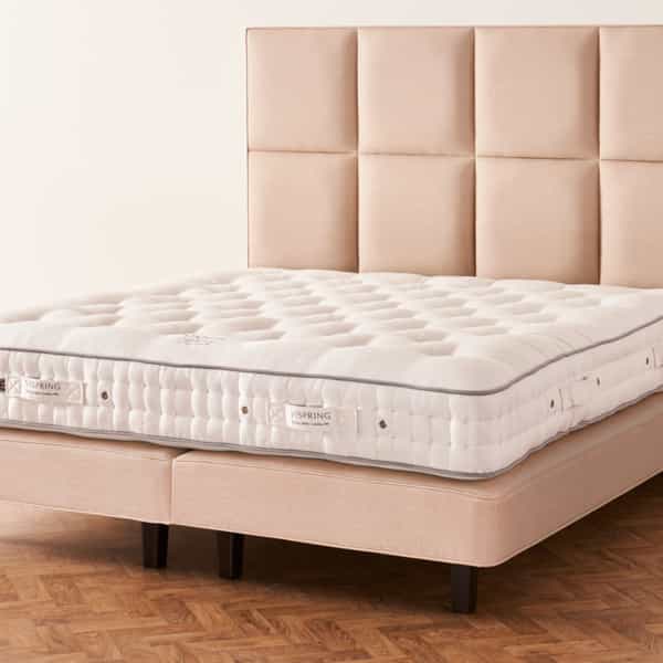 Elegant bedroom with a Vispring bed featuring a tufted upholstered headboard, plush mattress, and neutral bedding, placed beside a wooden nightstand with a lamp, in a room with wood-paneled walls, parquet flooring, and an open door letting in natural light.